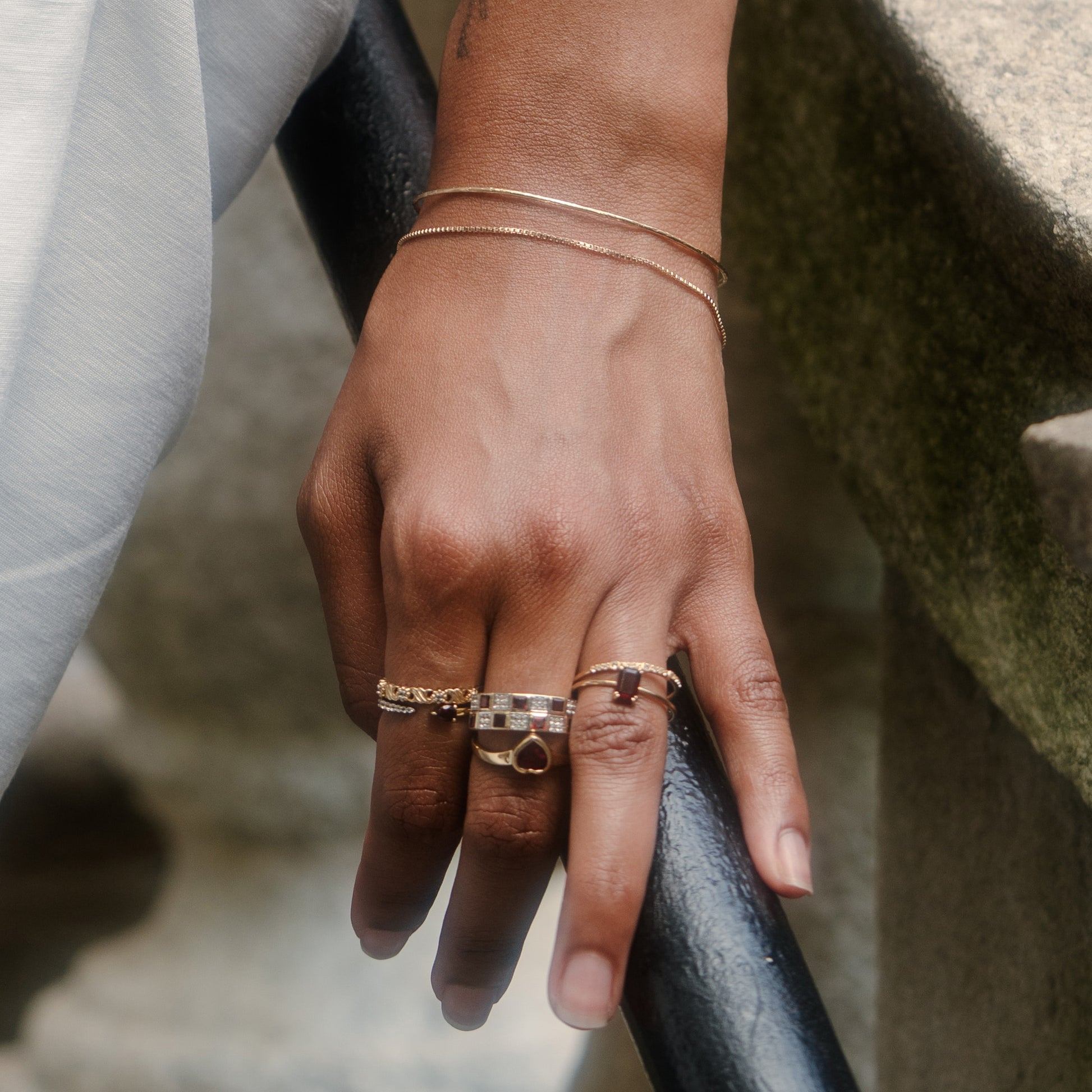 A close-up of a hand on a black railing, wearing the Solid Gold Delicate White Sapphire Stacking Band, assorted gold rings, and two gold bracelets. Stone surfaces and light-colored fabric are seen in the background.