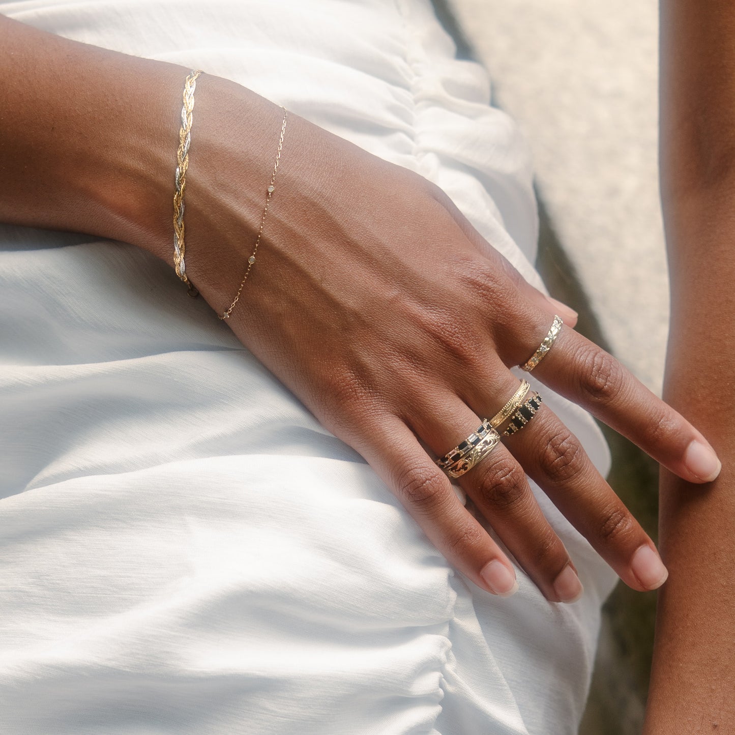 A close-up of a hand on a white ruched garment, adorned with delicate gold bracelets, several textured gold rings, and the Solid Gold Black and White Checkerboard Band.
