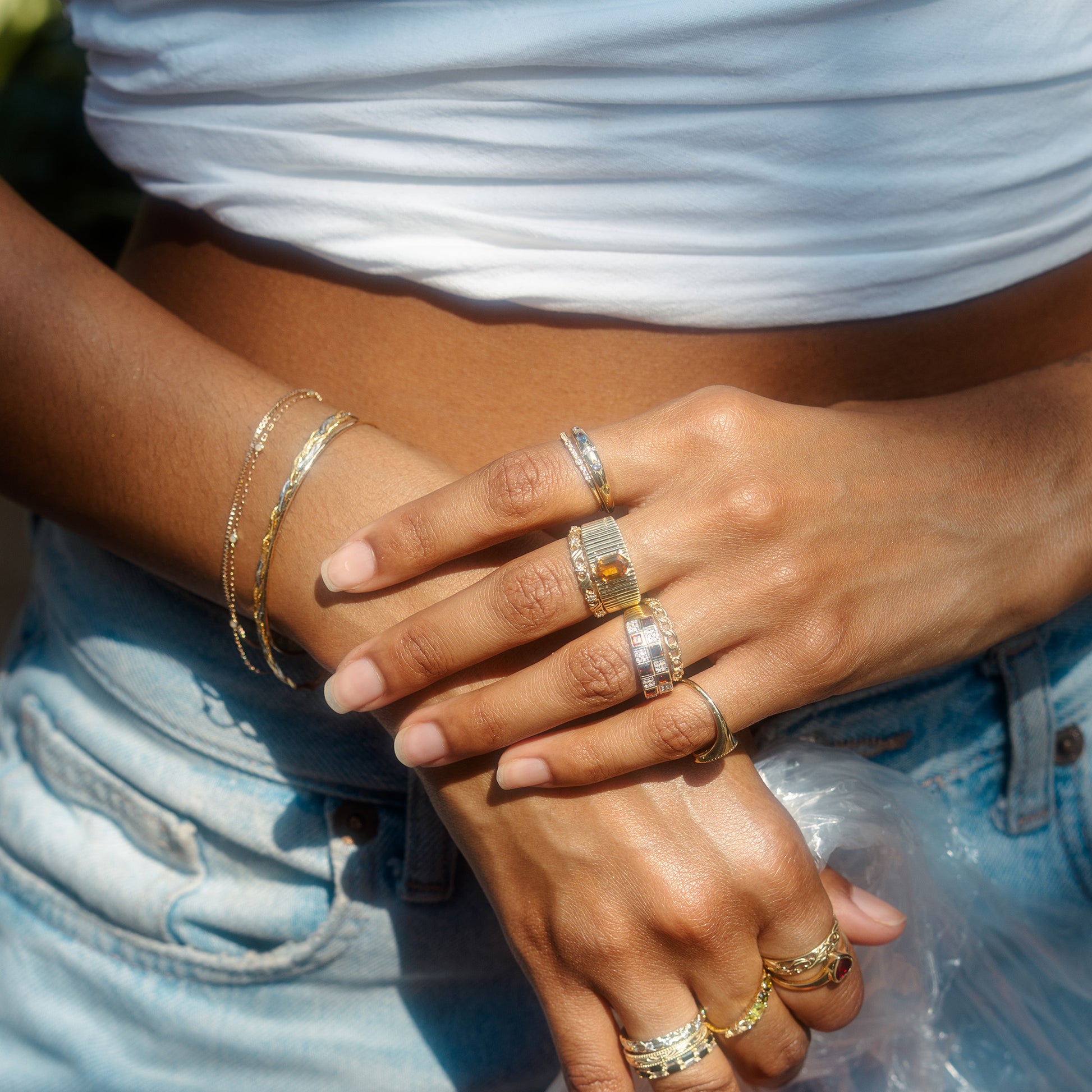 Wearing the Solid Gold Delicate White Sapphire Stacking Band and thin bracelets, a person in a white crop top and light blue jeans stands with fingers intertwined in natural sunlight.
