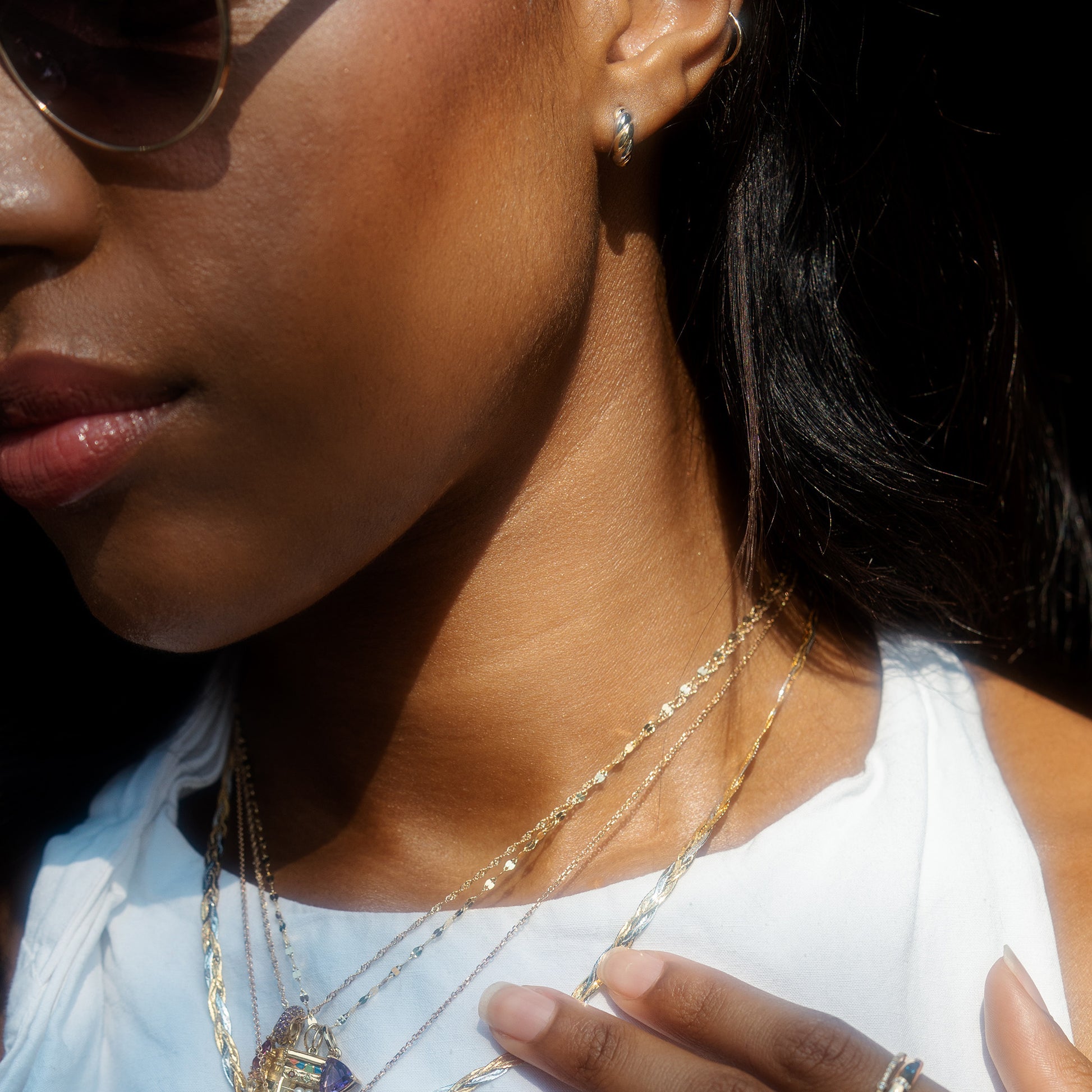 Close-up of a woman in sunglasses, a white top, and layered gold necklaces. She wears Solid Gold Mixed Metal Twisted Huggie Hoops, gently touching her chest to highlight the jewelry in natural light.