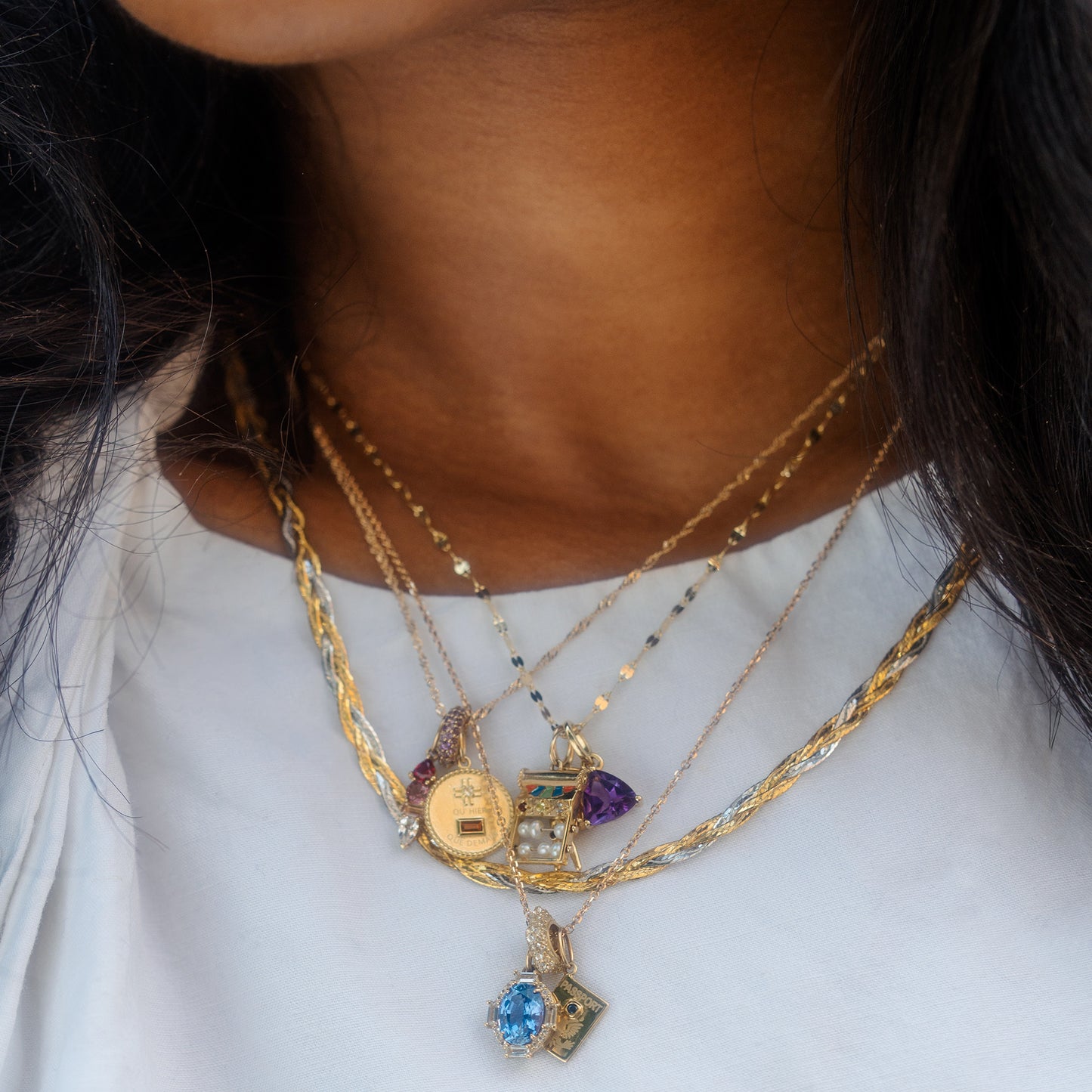 A woman wears a white shirt and several layered gold necklaces with milestone pendants, including the Solid Gold Anniversaire Charm, blue stone, purple gem, and gold coin. Her dark hair elegantly frames her neck.