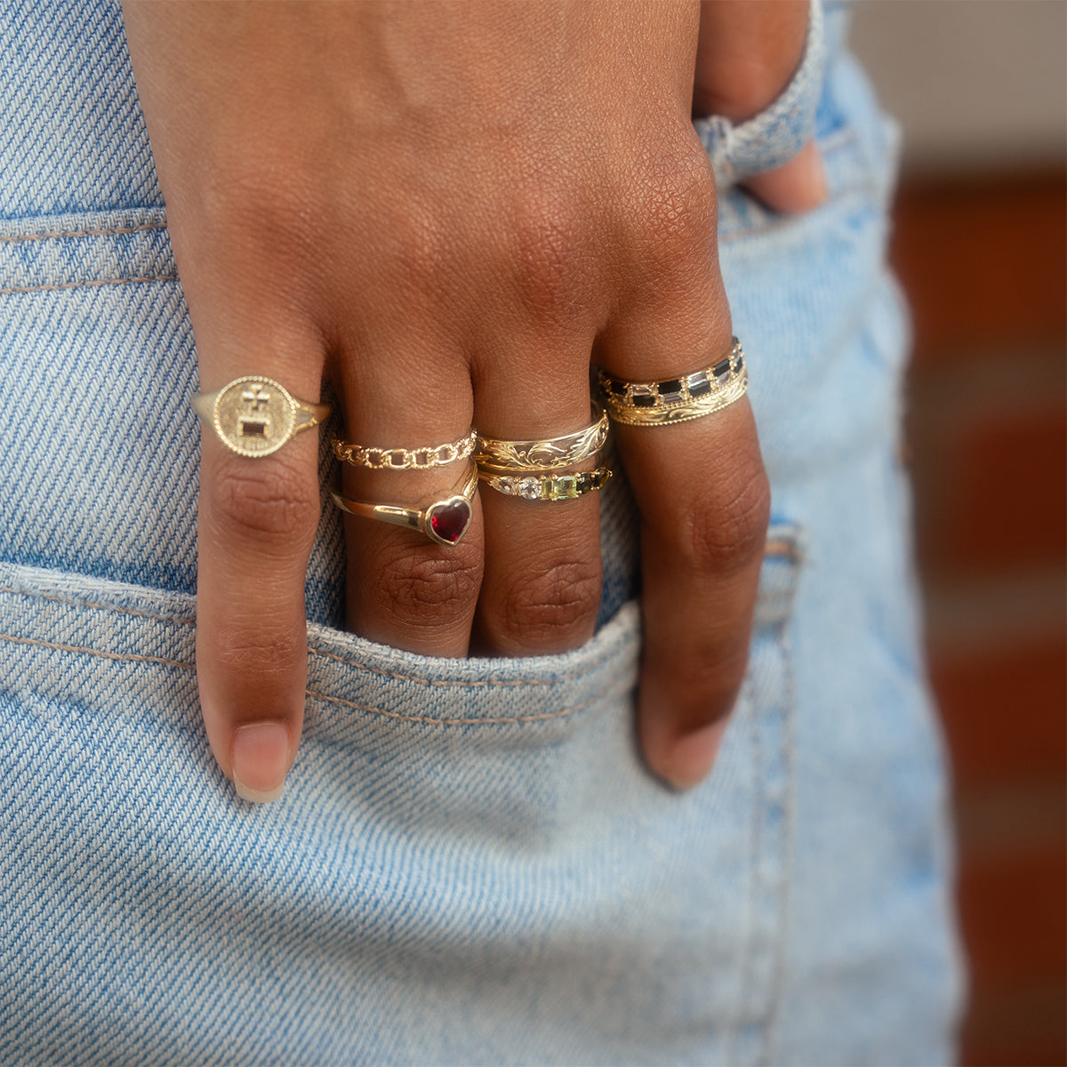 A close-up of a hand wearing five gold rings, including the Solid Gold Filigree Ring and one with a red heart, resting on the pocket of light blue denim jeans.