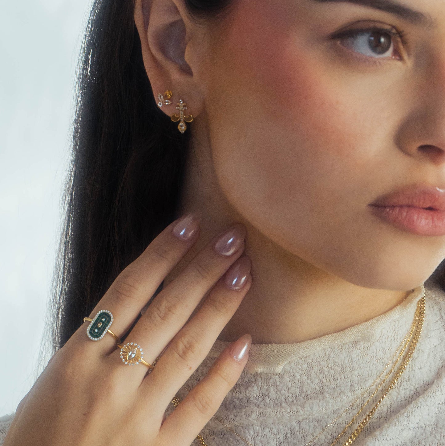A woman wears gold earrings, layered 14k yellow gold vermeil necklaces, and the Velvet Noir Navette Ring. Her manicured hand rests on her neck, and she is dressed in a light-colored top.