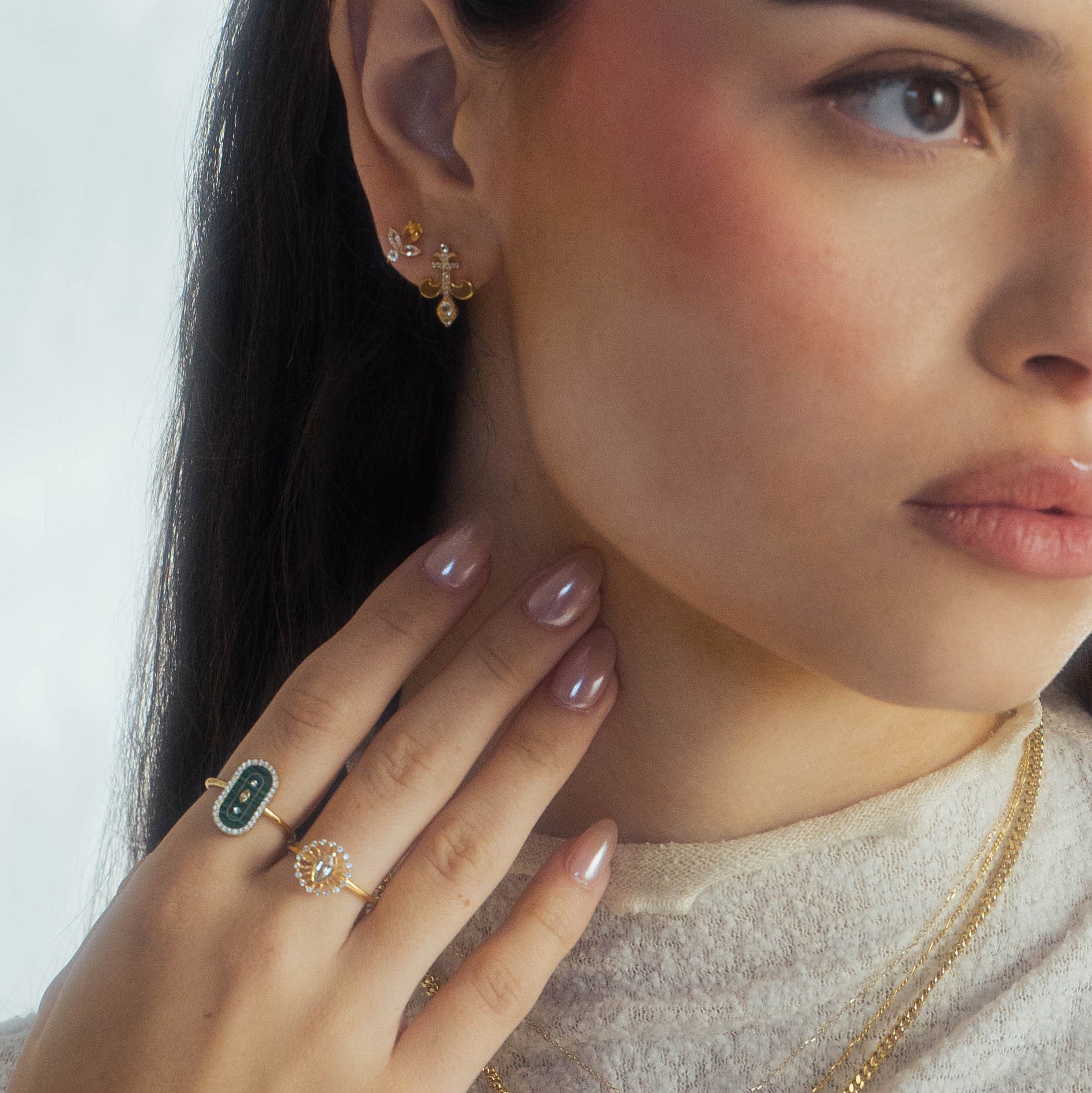 A woman wears gold earrings, layered 14k yellow gold vermeil necklaces, and the Velvet Noir Navette Ring. Her manicured hand rests on her neck, and she is dressed in a light-colored top.