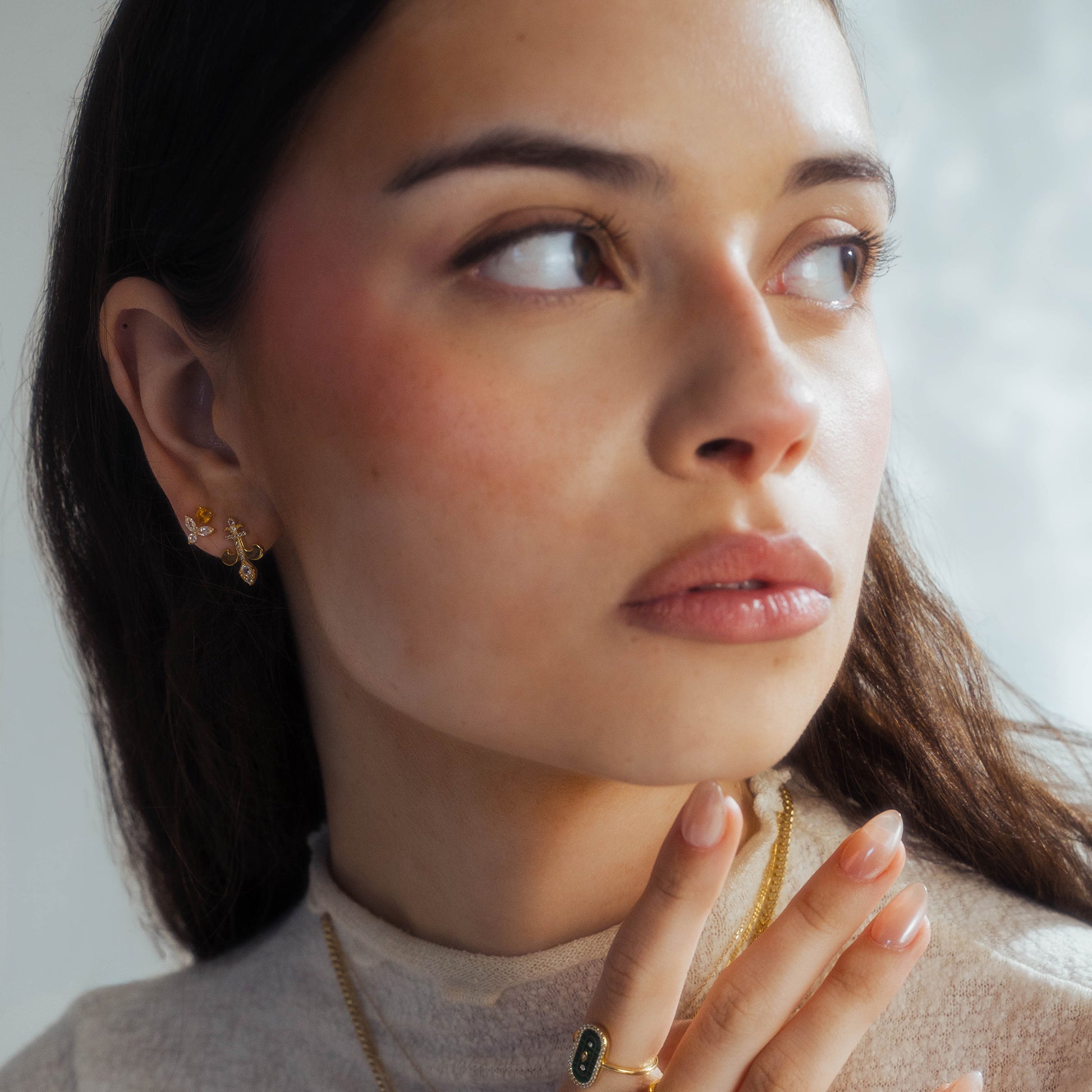 A woman with long brown hair and natural makeup gazes to the side, wearing Jazz Age Citrine Studs in 14k yellow gold vermeil. She pairs them with a gold necklace, a citrine and green stone ring, and a light beige top, her hand gently touching her neck.