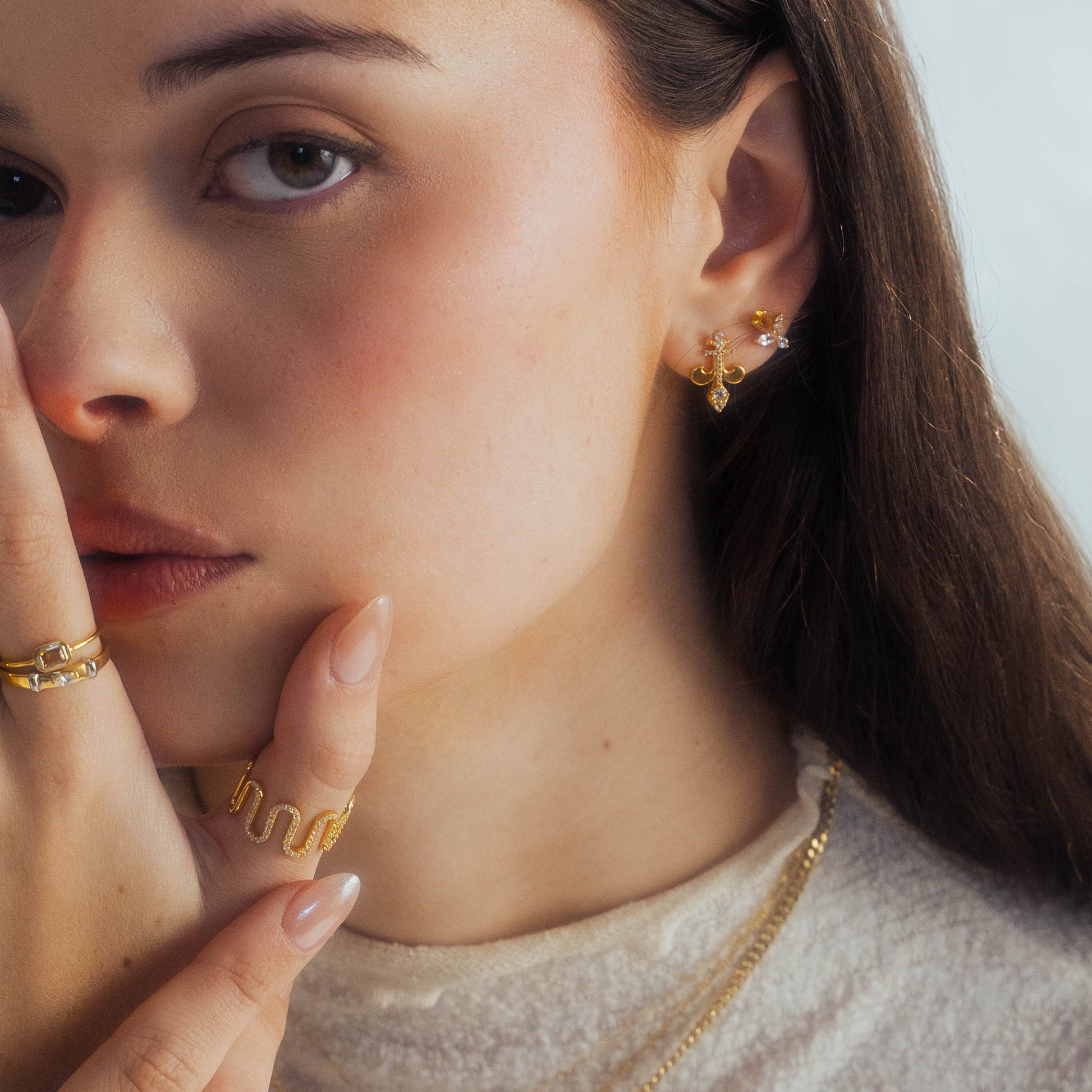 A woman with light skin and brown hair touches her face, showing off the Curtain Call Cascade Ring—14k yellow gold vermeil with sparkling white topaz. She has manicured nails and wears a soft beige top.