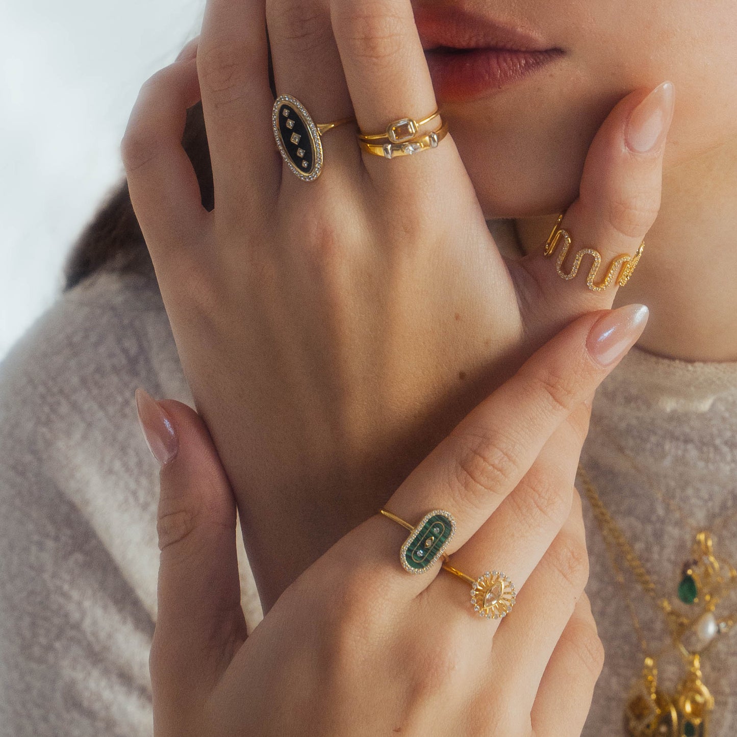 A close-up of a woman's hands adorned with gold rings, including the So Art Deco Cocktail Ring from Local Eclectic, paired with nude manicured nails and layered gold necklaces on her neck.