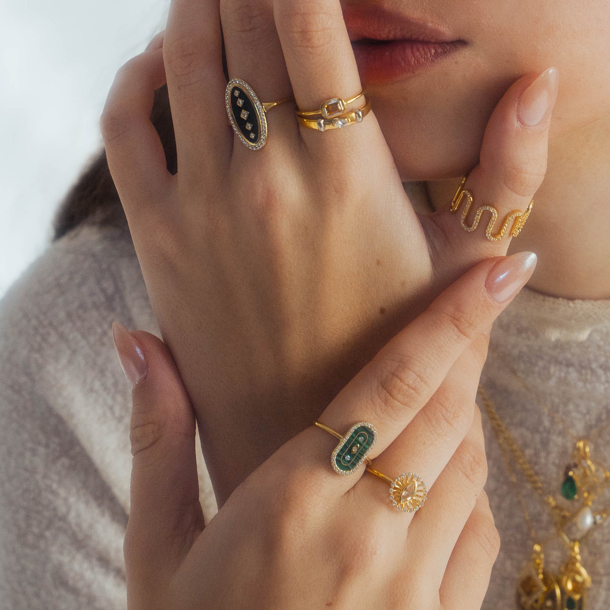 A close-up of a woman's hands adorned with gold rings, including the So Art Deco Cocktail Ring from Local Eclectic, paired with nude manicured nails and layered gold necklaces on her neck.
