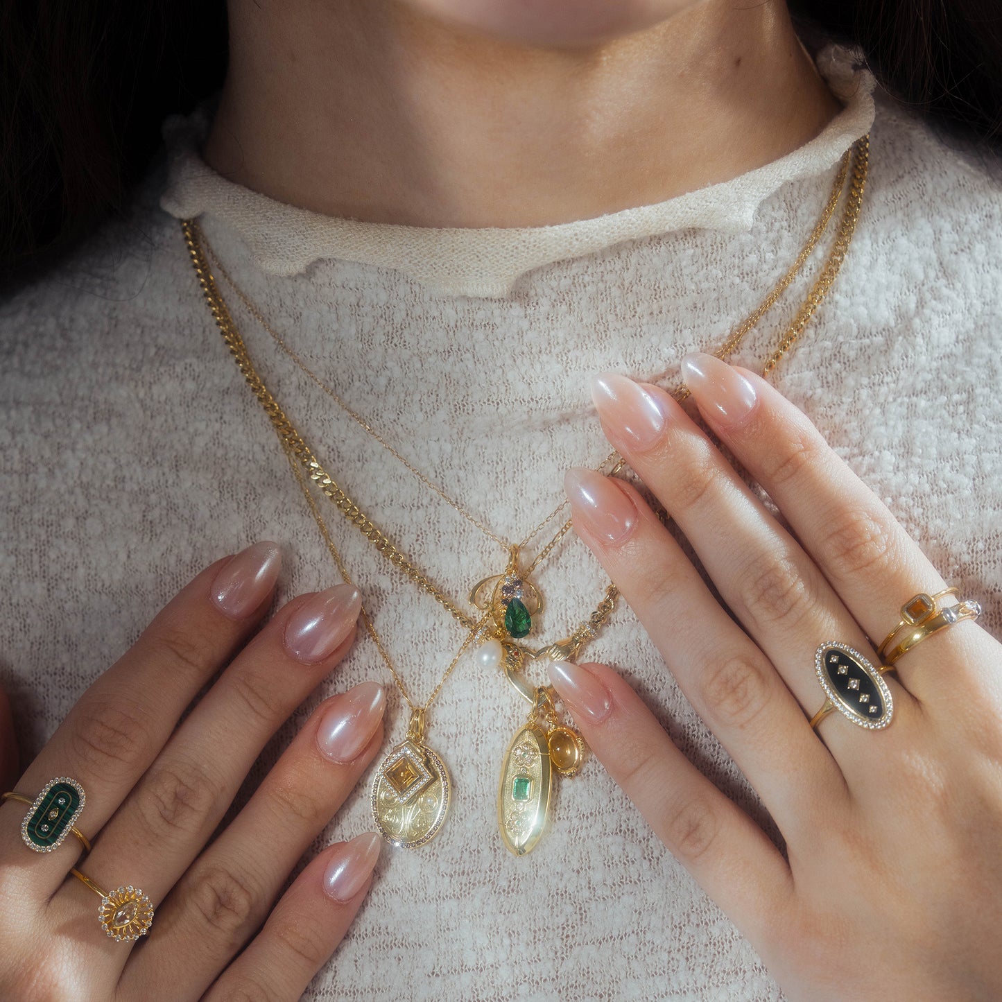 A close-up of a person wearing layered gold necklaces featuring the Vintage Inspired Pendant Charm and several rings, highlighting neatly manicured nude nails and a textured cream top.
