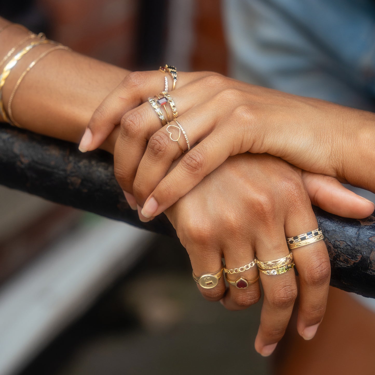 Two hands with manicured nails rest on a black railing, adorned with several rings and bracelets, including an everyday staple gold band and the Solid Gold Bezel Set Ruby Heart Ring. The background is softly blurred.