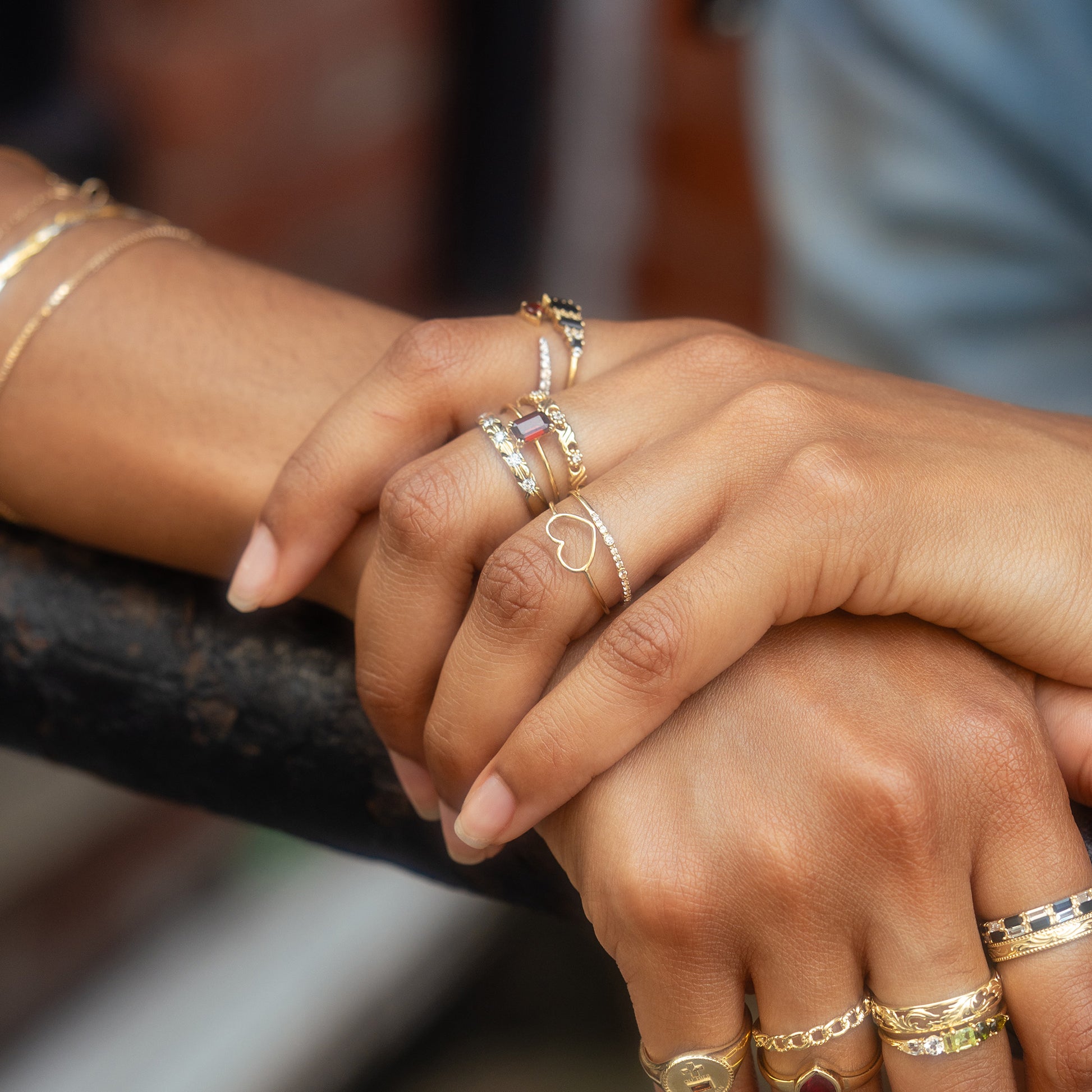 A close-up of two hands wearing multiple gold rings, including the Solid Gold Delicate Heart Ring, rests on a black metal railing. The blurred background highlights the elegance of the jewelry.