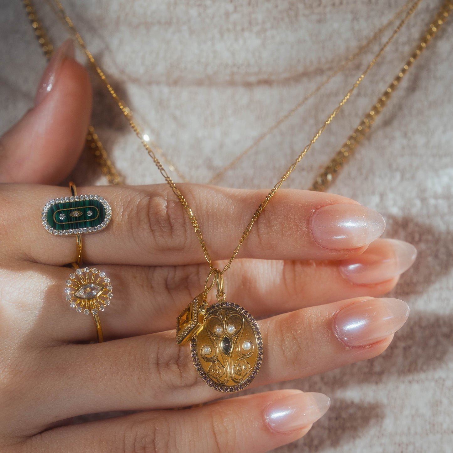 A close-up of a hand with long, glossy nails wearing two rings—one is the Silver Screen Starlet Ring, and the other green with diamonds—holding gold necklaces adorned with ornate gold and pearl pendants.