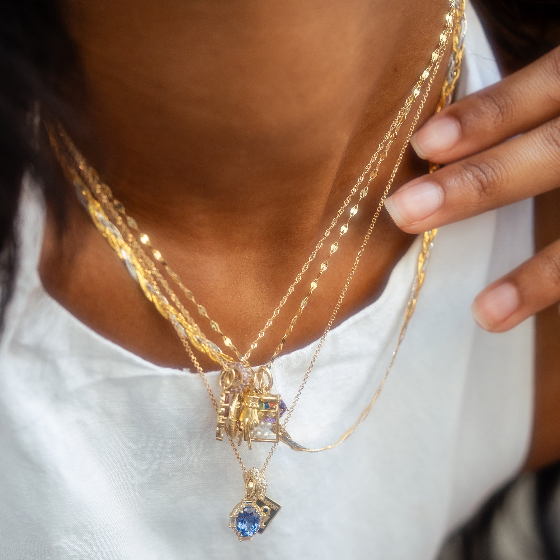 A close-up of a person in a white top, layering 9k solid yellow gold necklaces—including one blue gemstone pendant and the Solid Gold Body Language Charm—gently touching the charm with their hand.