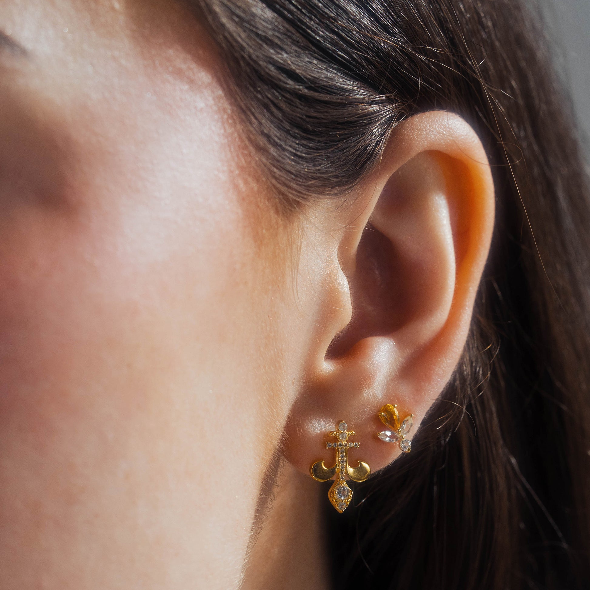 Close-up of a woman's ear with three piercings, including 14k yellow gold vermeil earrings: a fleur-de-lis with white topaz and two Jazz Age Citrine Studs featuring clear and citrine stones. Her brown hair is tucked behind her ear.