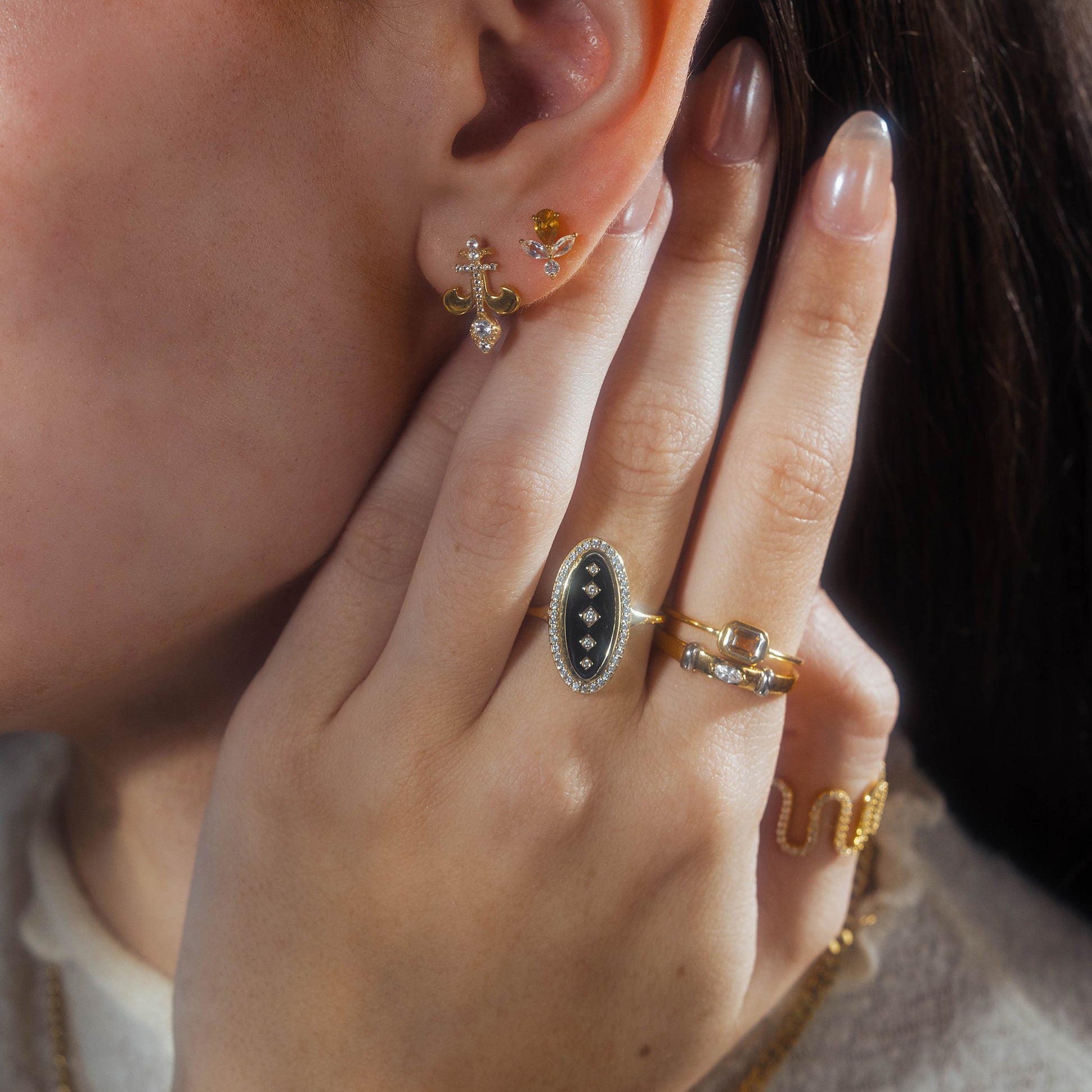 A close-up of a person’s ear and hand showcases multiple gold earrings, rings, and a chain bracelet, including the Velvet Noir Navette Ring with CZ black enamel. The person has long, manicured nails.