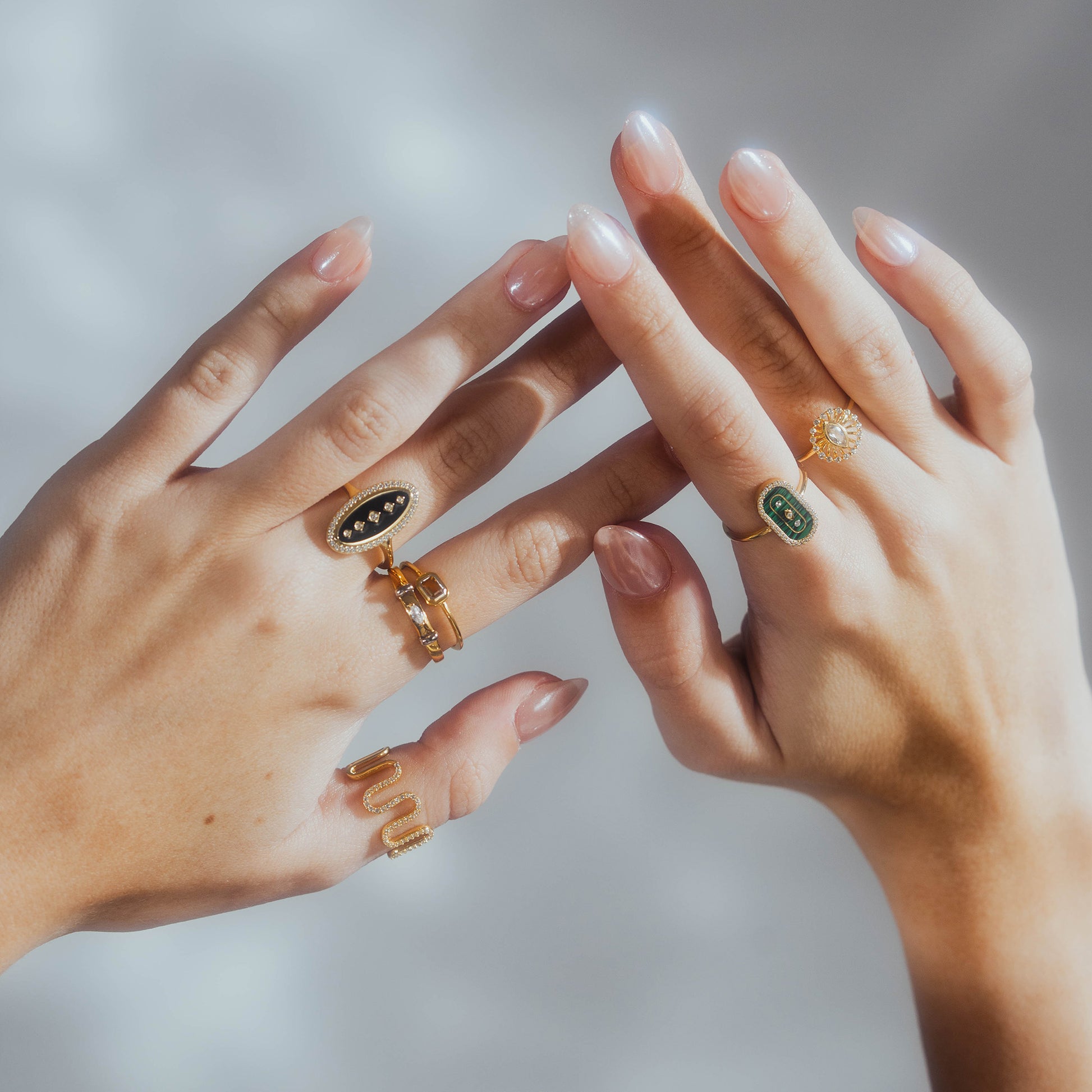 Two manicured hands display gold rings, including the Velvet Noir Navette Ring in 14k yellow gold vermeil and CZ black enamel designs, all set against a softly blurred background.