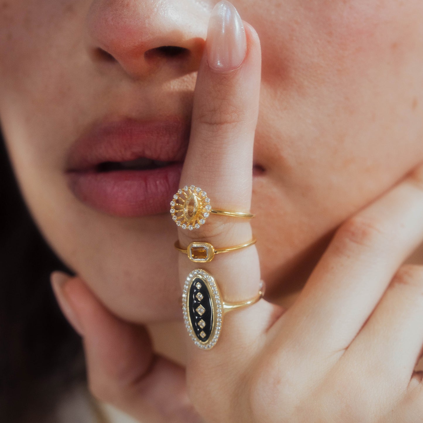 A person holds a finger to their lips, wearing the Velvet Noir Navette Ring and two others: a 14k yellow gold vermeil band with a clear rectangular gem and a ring with a round floral setting. Only their lower face and hand are visible.