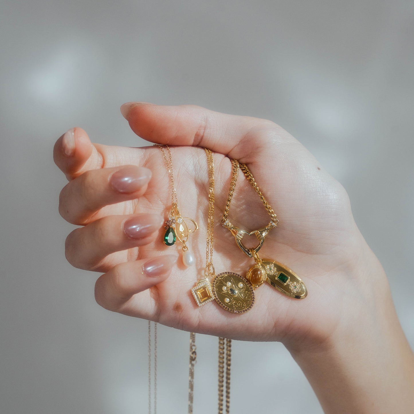 A hand with manicured nails holds several gold necklaces, each featuring a Vintage Inspired Pendant Charm—such as a gemstone, coin, and pearl—set against a soft, light background.