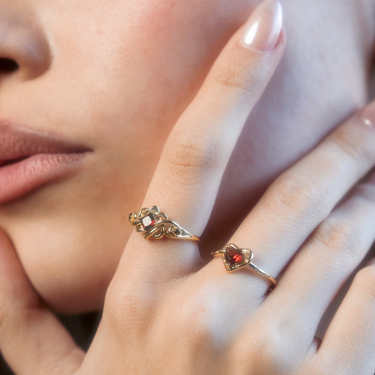 A close-up of a person's face and hand wearing two gold rings: a garnet heart ring in 14k yellow gold vermeil and the Lover's Knot Heart Ring with an intricate geometric design. The person has smooth skin and pale pink lips.