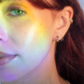 Close-up of a woman’s face with a rainbow light across her cheek, green eye, and brown hair. She wears Solid Gold Black Diamond Open Ear Huggers and has a soft, natural makeup look.