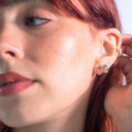 A close-up of a fair-skinned woman with red hair wearing Solid Gold Ombre Birthstone Huggie Hoops and a gold ear cuff on her left ear. Her hand gently touches her ear, highlighting the colorful hoops. She wears natural makeup.