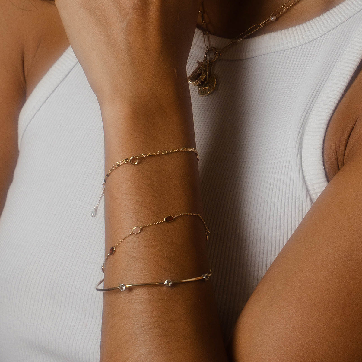 A person showcases the Solid Gold Geometric Glimmer Mixed Metal Bracelet, styled with layered necklaces from Local Eclectic's Family Gold collection, while wearing a white ribbed tank top and resting their hand near their face. The focus is on the jewelry.