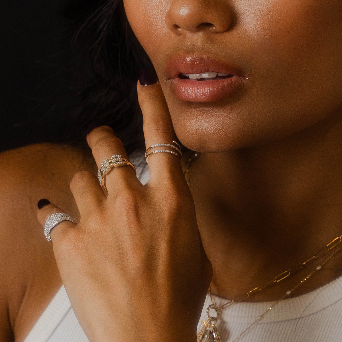 A close-up of a woman wearing the Solid Gold Mirrored Light Diamond Statement Ring and a white sleeveless top, with her hand gently touching her chin against a dark background.