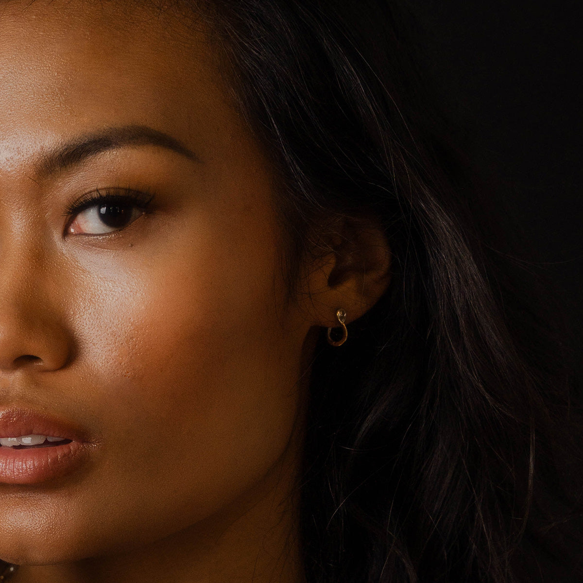 Close-up of a woman’s face with smooth skin and long dark hair against a dark background, revealing one eye, eyebrow, cheek, and part of her lips. She wears the Solid Gold Mixed Metal Duo Huggies from Local Eclectic.