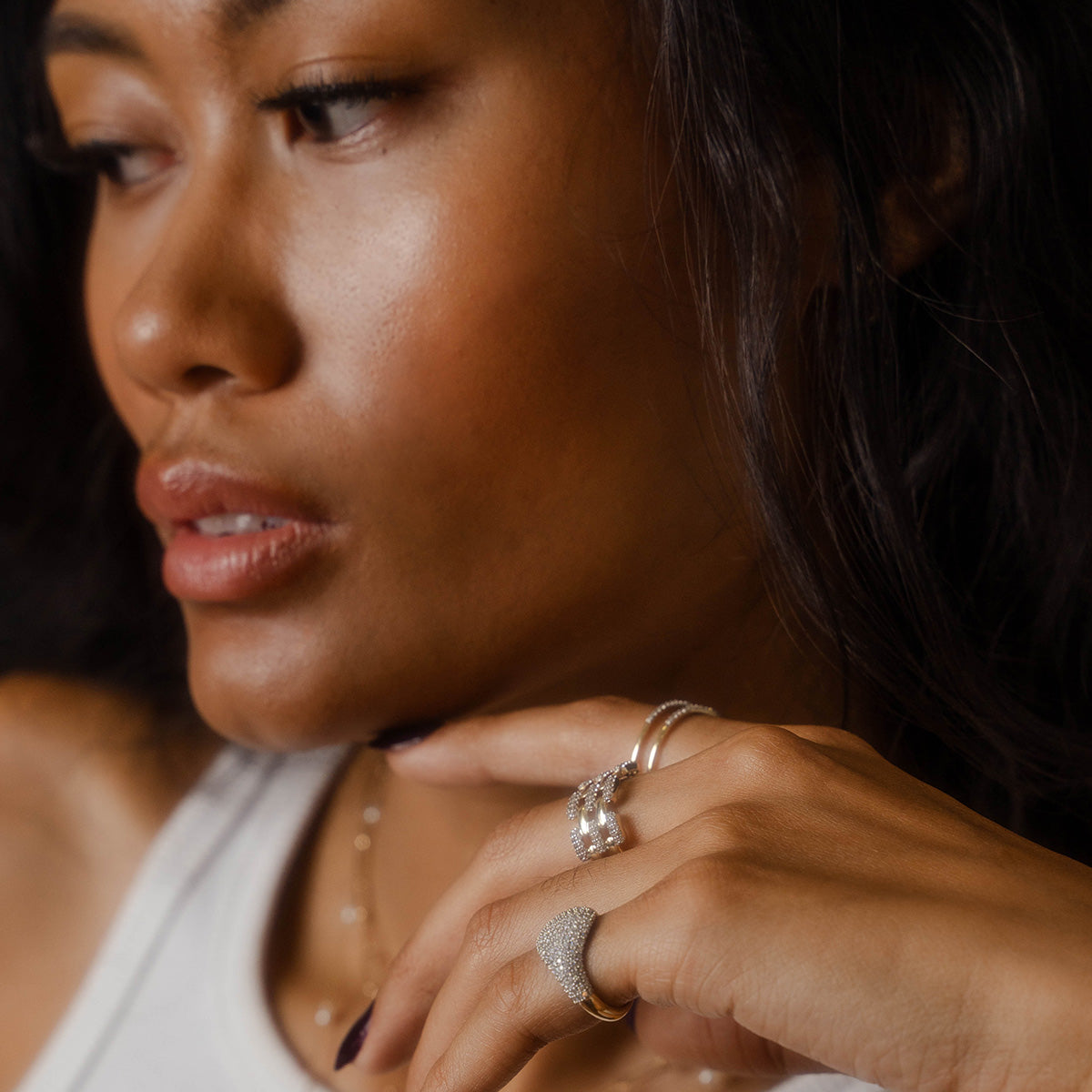 A woman with long dark hair gazes to the side, wearing a white top and the Solid Gold Mirrored Light Diamond Statement Ring from Local Eclectic’s Family Gold collection. Soft, warm lighting highlights her radiant skin.