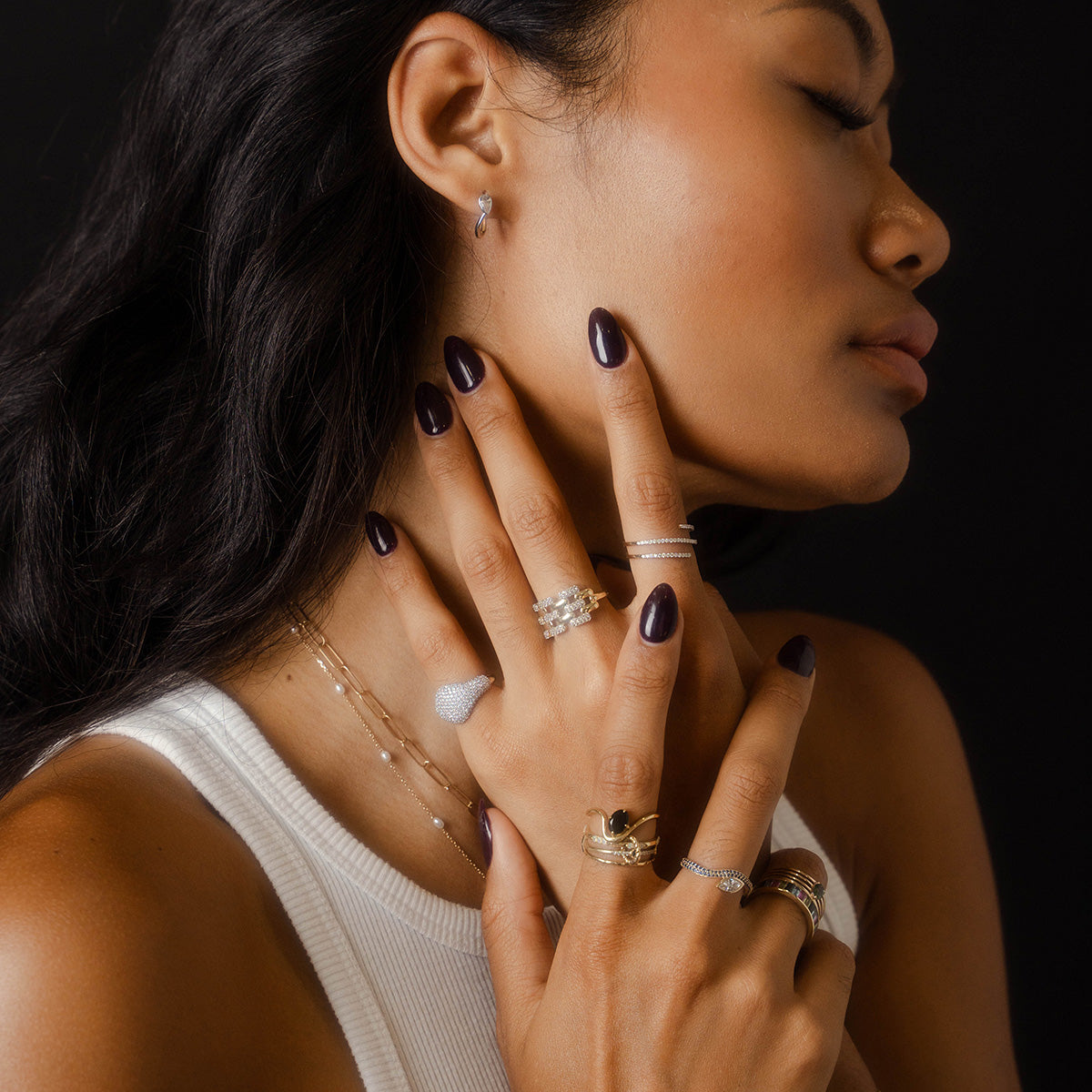 A woman poses in profile against a dark background, her dark polished nails grazing her neck. She wears the Solid Gold Mirrored Light Diamond Statement Ring, its shine adding elegance to her look.