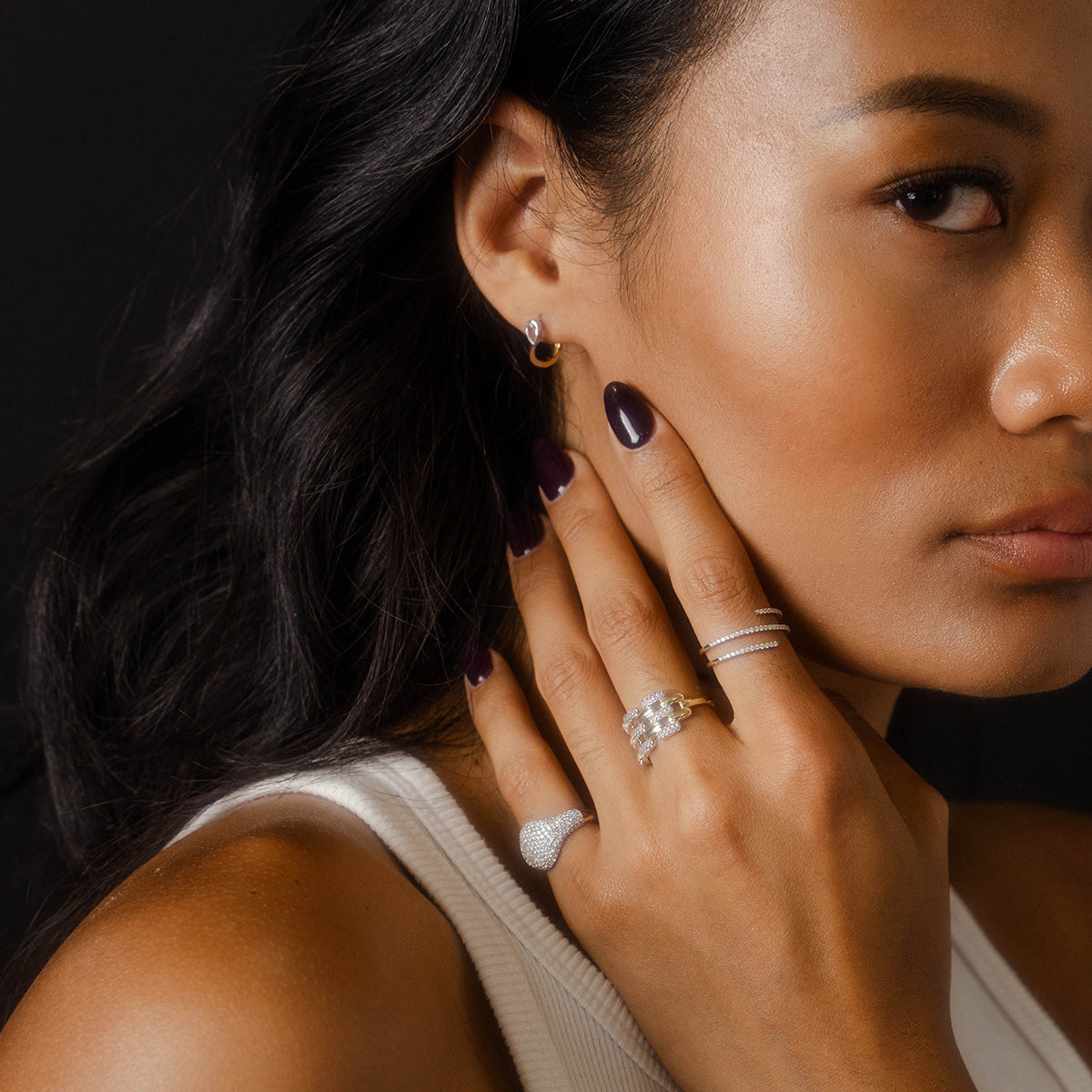 A woman with long dark hair and dark nail polish, wearing a white top, elegantly showcases the Solid Gold Mirrored Light Diamond Statement Ring from Family Gold as she poses with her hand on her face against a black background.