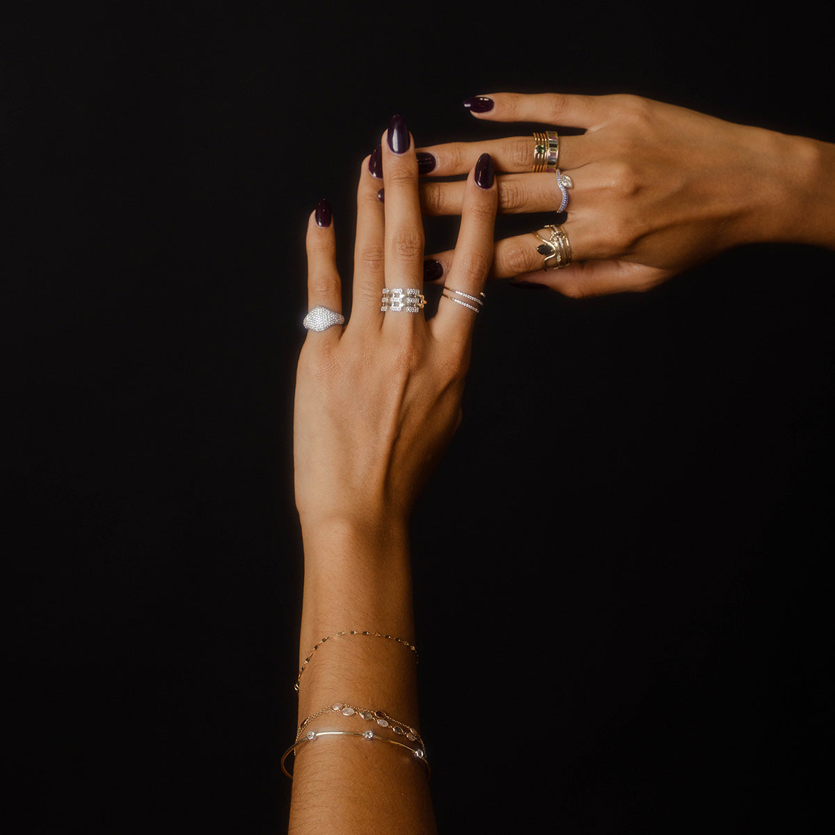 Two hands with dark nail polish display elegant rings and bracelets against a black background. Featured is the Solid Gold Mirrored Light Diamond Statement Ring, adding a stylish, sparkling touch to the layered gold and silver jewelry.