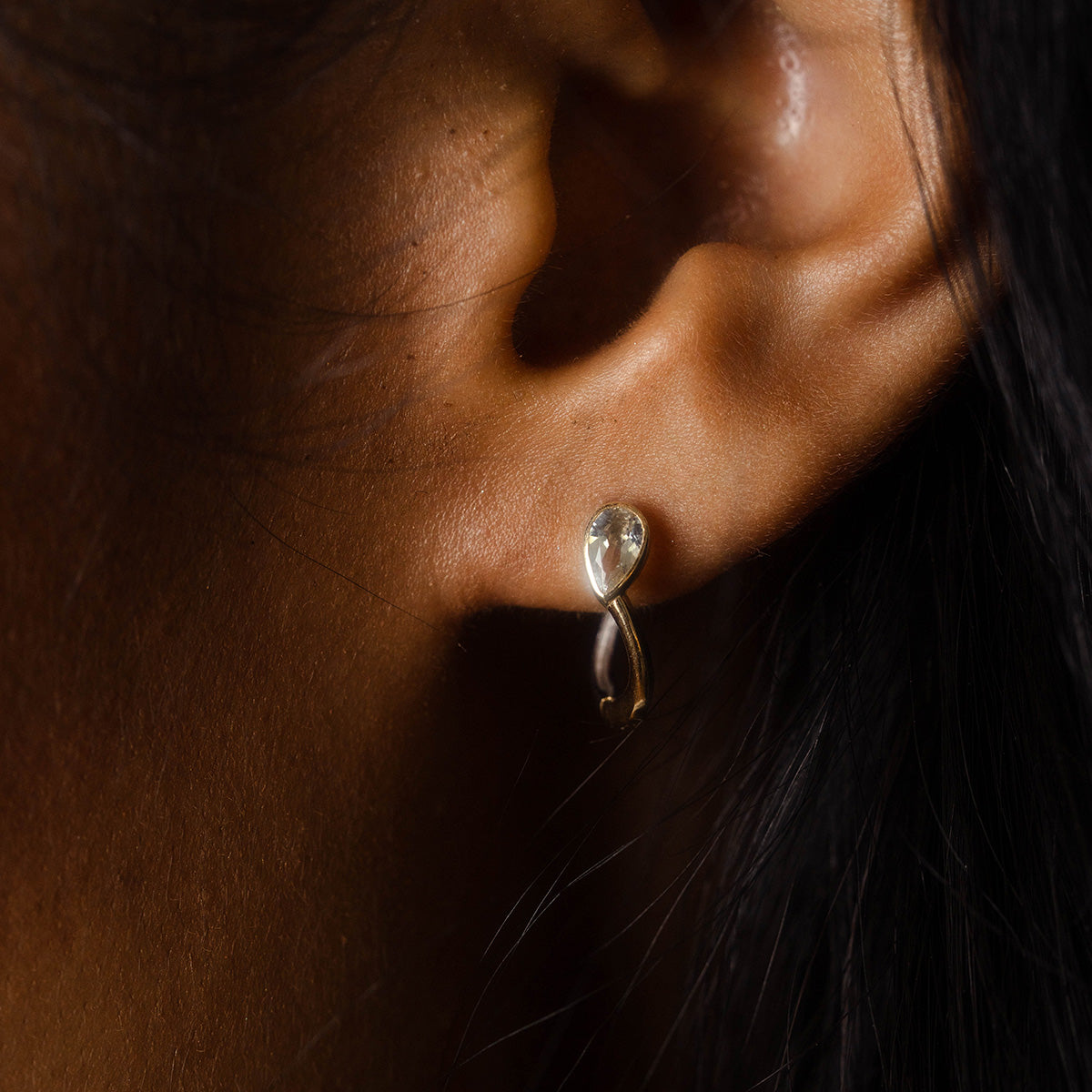 Close-up of a person's ear wearing Solid Gold Mixed Metal Duo Huggies from Family Gold at Local Eclectic, set against dark hair and warm brown skin.
