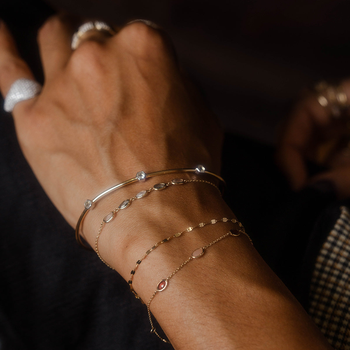 A close-up of a hand wearing several Solid Gold Geometric Glimmer Mixed Metal Bracelets with shimmering accents, set against a dark background. Family Gold rings on the fingers complete the elegant, minimalistic look.