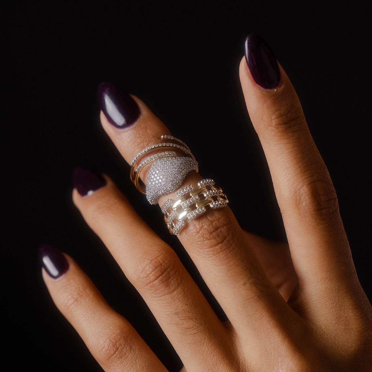 A hand with dark purple polished nails wears Local Eclectic’s Solid Gold Shifting Shimmer Diamond Wrap Ring on the middle finger, sparkling against a black background.
