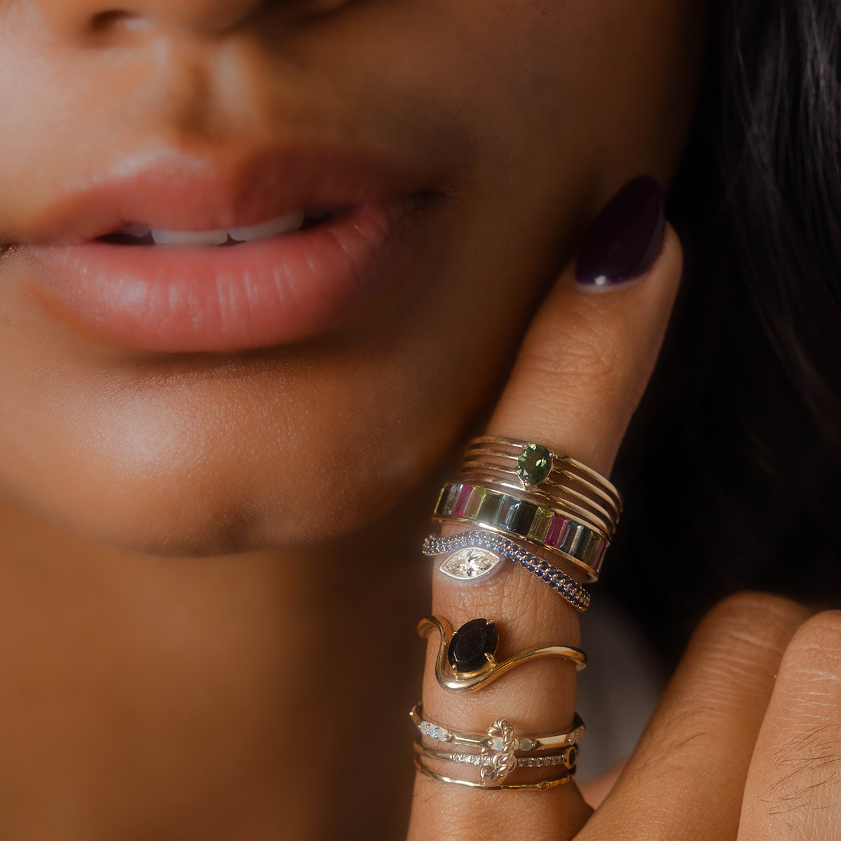 A close-up of smooth lips and fingers with dark nail polish, featuring the Solid Gold Nesting Gleam Sapphire and Diamond Ring from Local Eclectic, set with sparkling sapphires and diamonds.