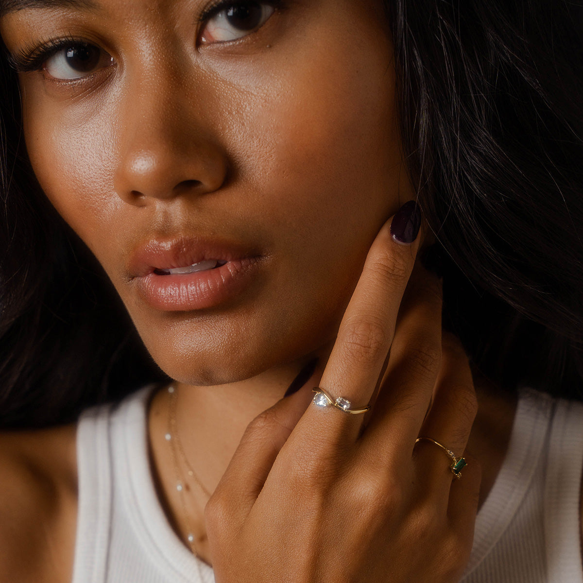 A woman with long dark hair and glowing skin wears a white sleeveless top. She touches her face, highlighting the 2026 April Capsule Floating Duo White Topaz Ring and a delicate necklace.