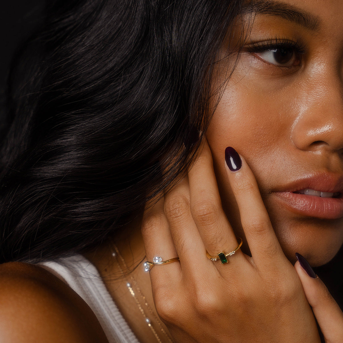 A close-up of a woman with dark hair touches her face, wearing the 2026 May Capsule Delicate Emerald Solitaire Ring and gemstone rings. She has smooth skin, natural makeup, dark nails, a white top, and layered necklaces.