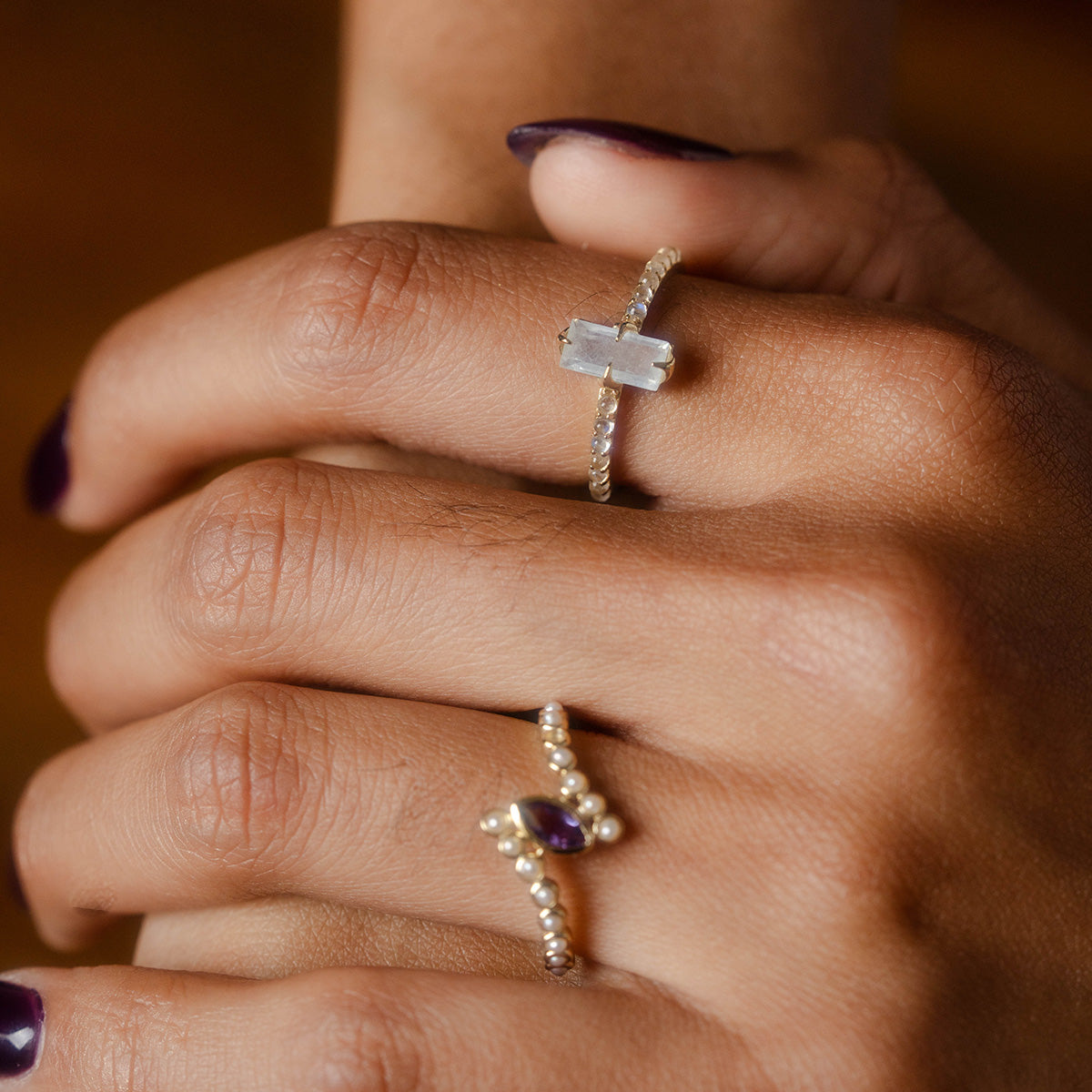 Close-up of hands with dark purple nail polish, wearing two rings: a yellow gold vermeil ring with a rectangular clear stone and beaded band on the middle finger, and the 2026 March Capsule Delicate Aquamarine Solitaire Ring on the ring finger.