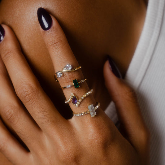 A close-up of a hand with dark purple nail polish wearing several gold rings, including the 2026 May Capsule Delicate Emerald Solitaire Ring, resting gently on a bare shoulder.