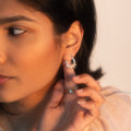 A woman with medium skin tone holds her ear, displaying the Carved Moon Mother of Pearl Prophecy Earrings. She also wears a gold ring and a peach top, with soft natural light highlighting her face.