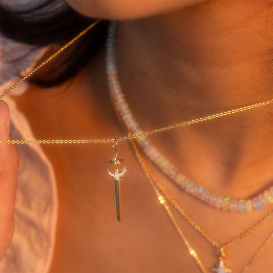 A close-up of a person holding the Buffy Dagger Necklace—a gold, sword-shaped pendant. They wear layered gold chains and a beaded choker, with their softly lit lips and neck visible in warm light.