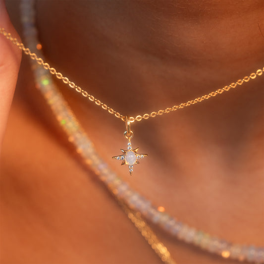 A close-up of a person wearing the Sabrina Opal Portal Necklace—a gold chain with a star-shaped white gemstone pendant—layered with a beaded necklace. Warm light highlights the jewelry and skin.