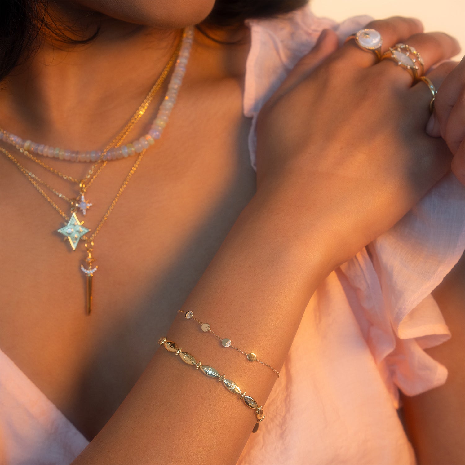 A woman in a pale pink top wears the Astral Guide Amulet Necklace along with layered gold jewelry featuring gemstones and unique charms, touching her shoulder gently.