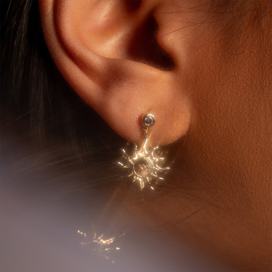 Close-up of a person's ear adorned with the Summer Solstice Earrings—gold sun-shaped design featuring a small central gemstone, hanging below a simple stud. Lighting enhances the earrings' shiny and intricate details.