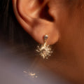 Close-up of a person's ear adorned with the Summer Solstice Earrings—gold sun-shaped design featuring a small central gemstone, hanging below a simple stud. Lighting enhances the earrings' shiny and intricate details.