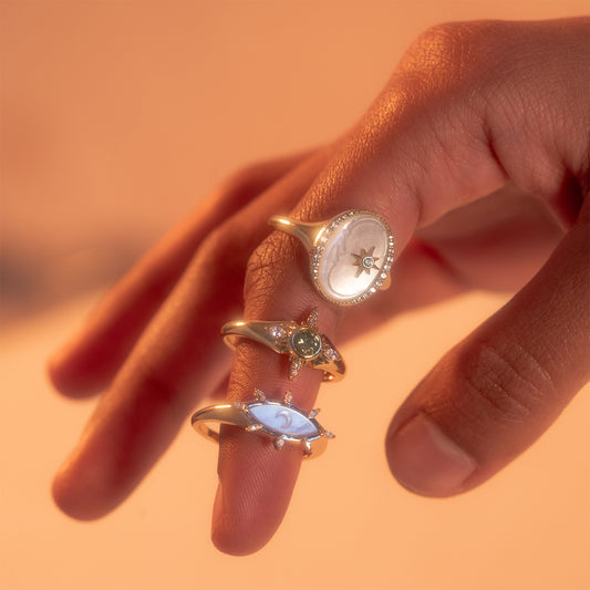 A hand with three fingers extended, each wearing a unique silver ring—including the Carved Moon Mother of Pearl Divination Ring—featuring different gemstone designs, shown in warm, soft lighting.