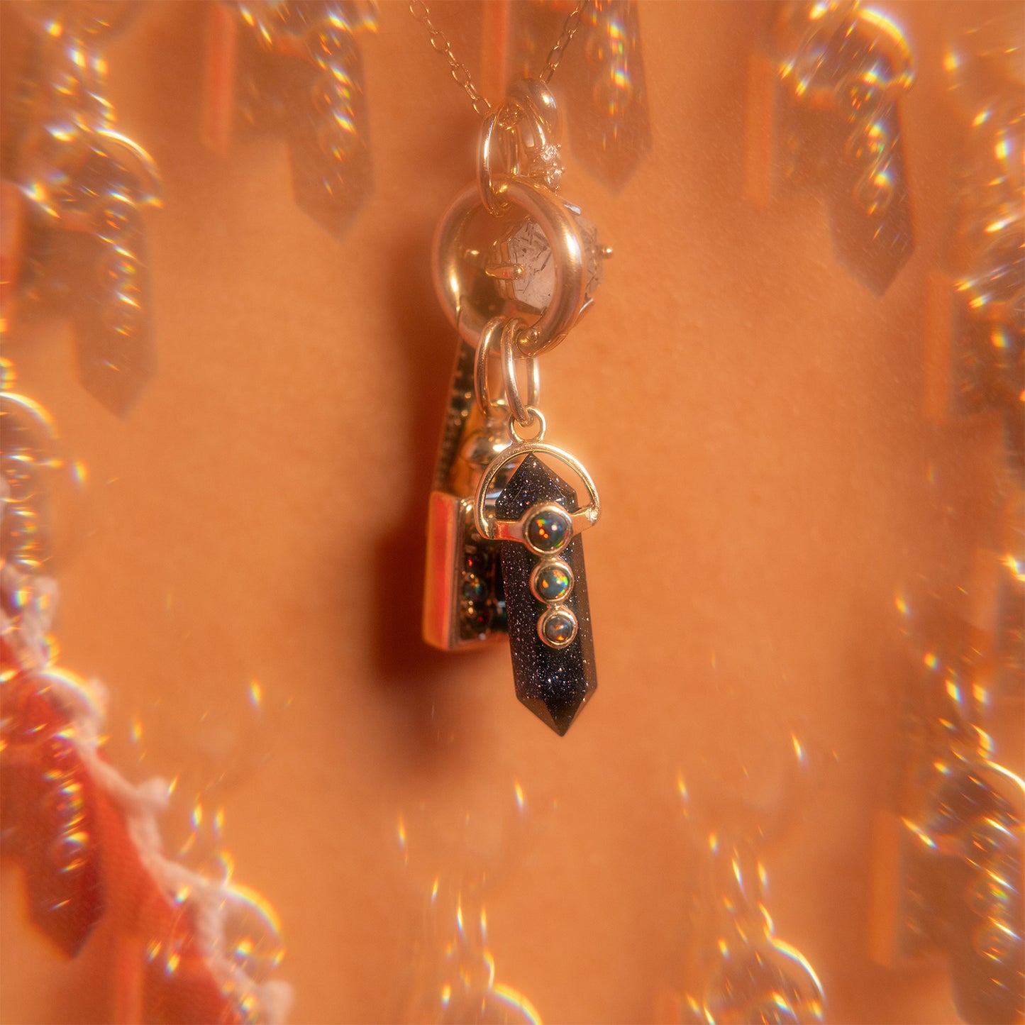 A close-up of the Solid Gold Amulet Charm featuring a dark crystal pendant with three small gold spheres, set against a warm, kaleidoscopic background—a true Local Eclectic exclusive.
