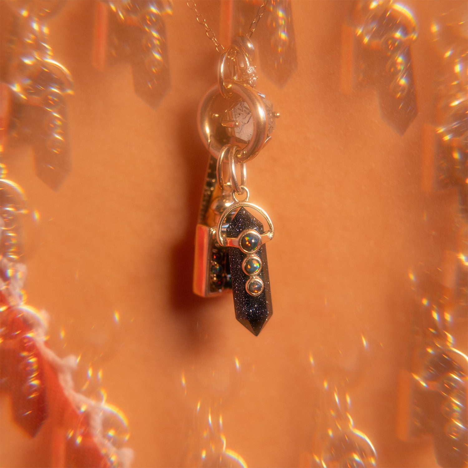 A close-up of the Solid Gold Amulet Charm featuring a dark crystal pendant with three small gold spheres, set against a warm, kaleidoscopic background—a true Local Eclectic exclusive.
