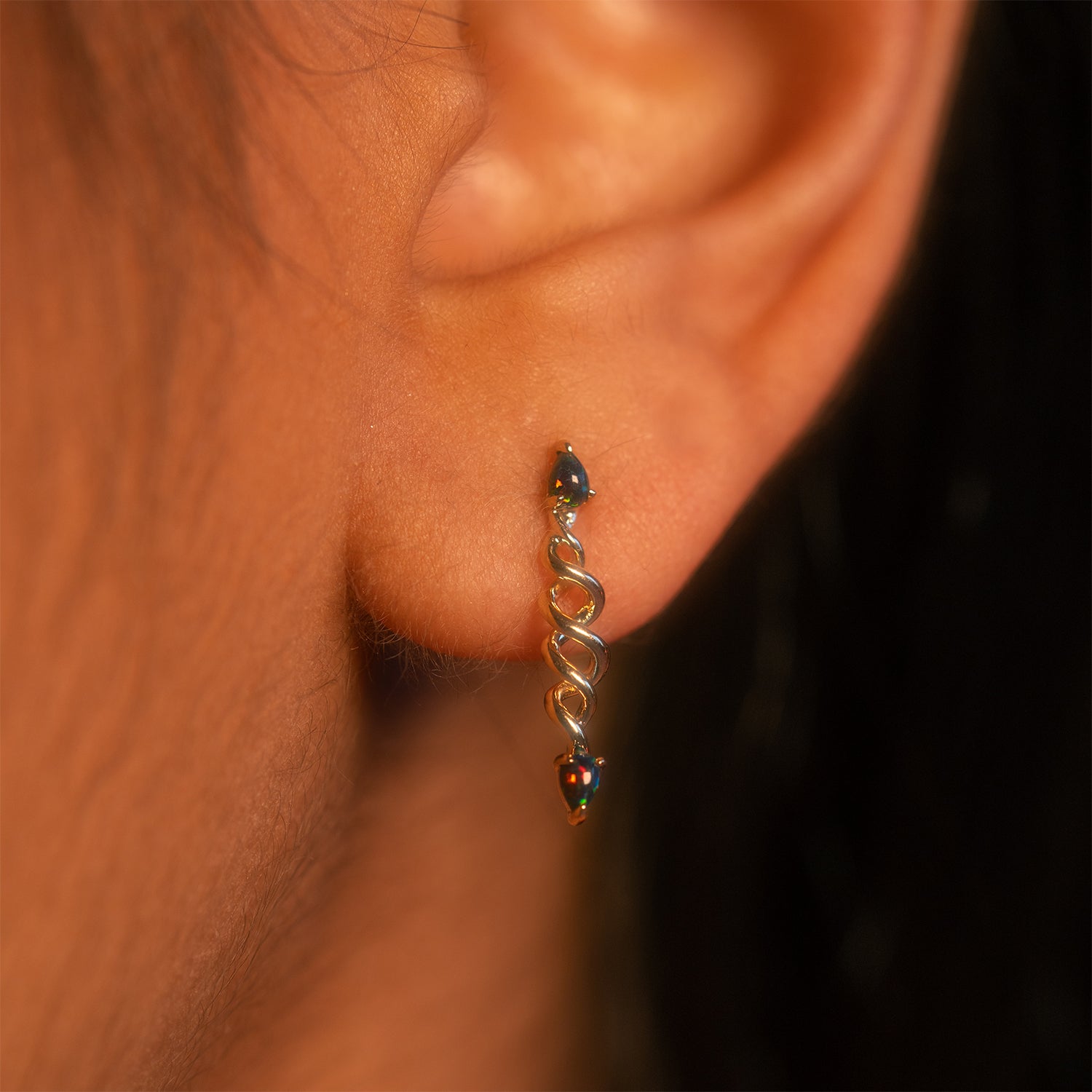 Close-up of a person's ear wearing the Solid Gold Black Opal Cosmic Spiral Earring, featuring an elegant intertwined gold spiral with small black opal teardrop stones at each end.